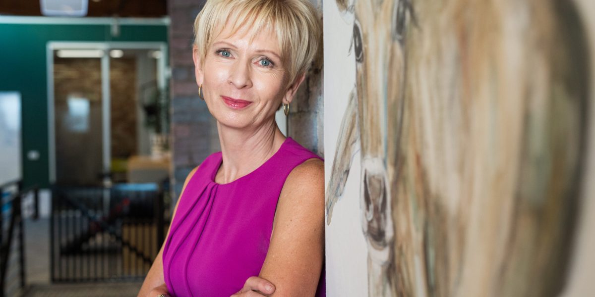 Lyn White leaning against a wall with arms crossed, looking at camera.