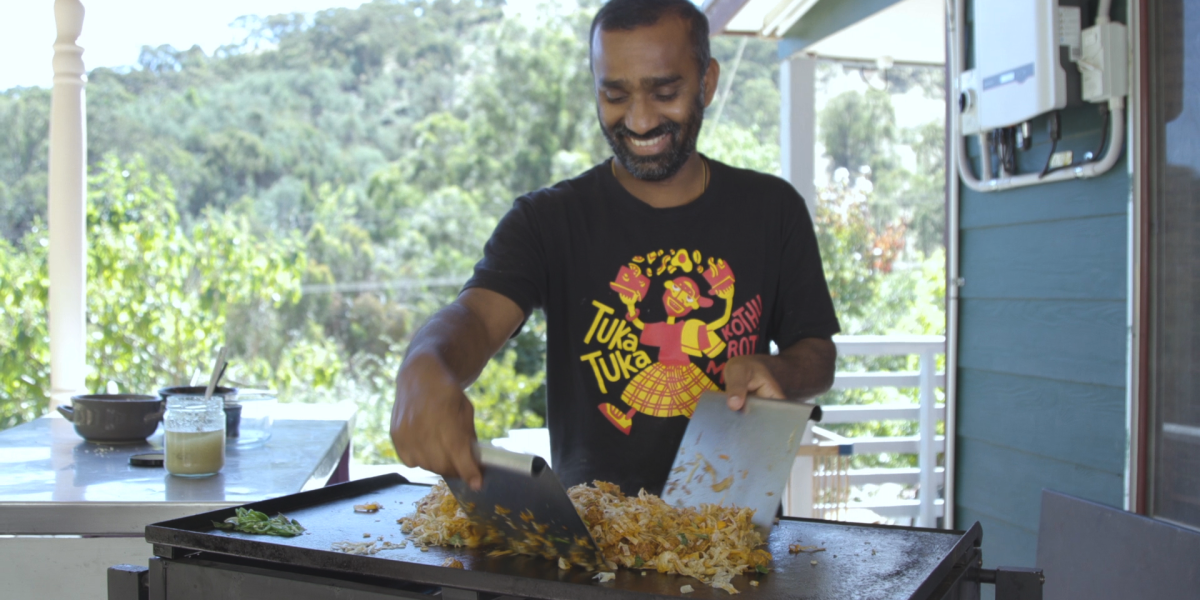 Niro is cooking a big batch of Kothu roti on a large grill in an outdoor setting, trees in the background.