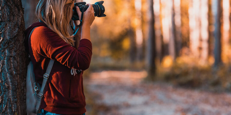 A photographer with long hair wearing backpack taking photo while standing close to a tree