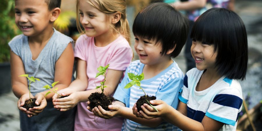 Four young kids with big grins hold small plants in handfuls of soil.