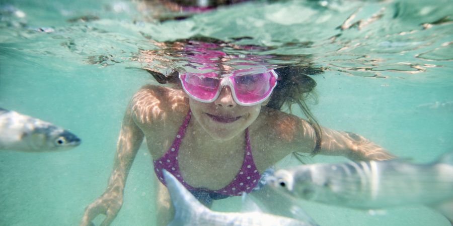 A girl in pink goggles smiles underwater surrounded by silver fish.