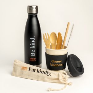 Black drink bottle with 'Be kind' and the AA logo, next to reusable coffee cup with 'Choose kindness' printed on the black band in the middle of the cup. Reusable bamboo cutlery set with straw and straw cleaner is sitting in the coffee cup and the carry pouch for the set is lying in front. Pouch says 'Eat kindly' and has the AA logo printed on it