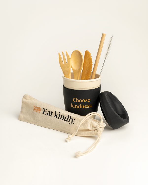 Reusable coffee cup with 'Choose kindness' printed on the black band in the middle of the cup. Reusable bamboo cutlery set with straw and straw cleaner is sitting in the coffee cup and the carry pouch for the set is lying in front. The lid for the cup is leaning against the cup. Pouch says 'Eat kindly' and has the AA logo printed on it