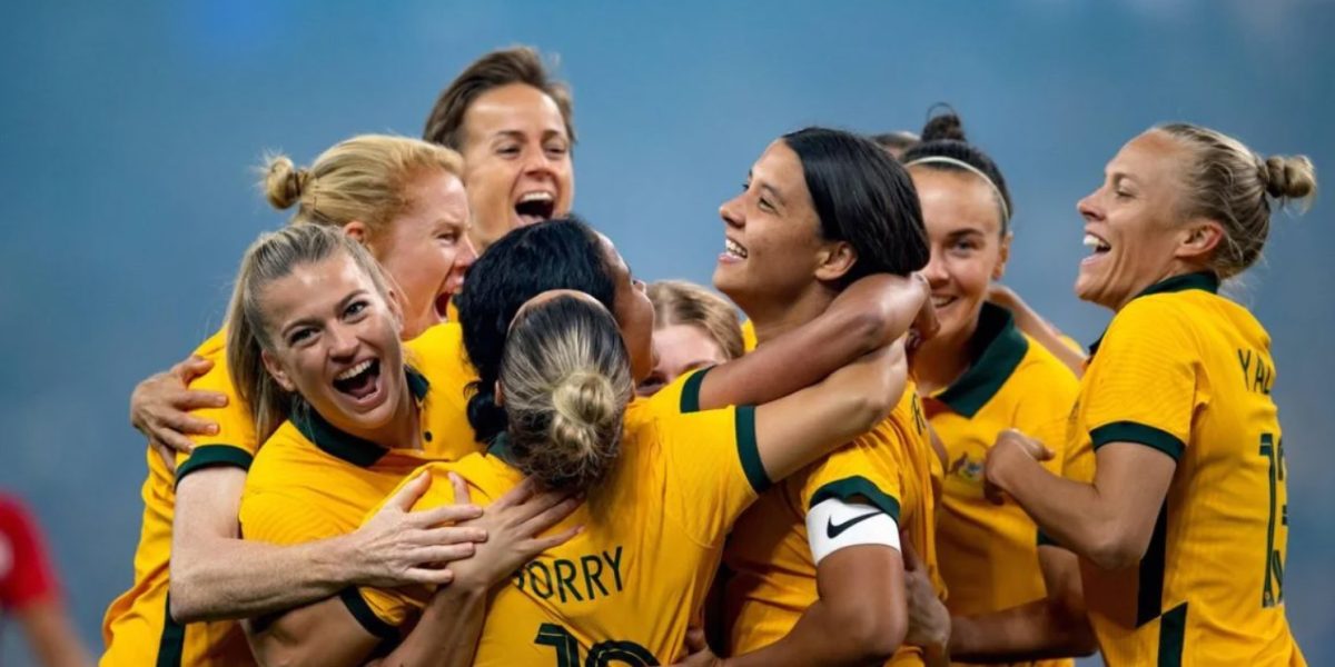 Seven players for the Matildas celebrate on the field. They are wearing yellow jerseys and are all smiling and hugging.