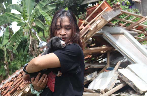 A woman holds a rescued puppy in front of a collapsed building.