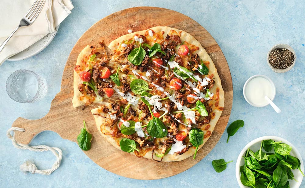 Overhead view of a Turkish-style pizza on a wooden serving board. The pizza is made using the Birds Eye plant-based lamb strips. Several condiment dishes sit on the blue bench around it with added ingredients.