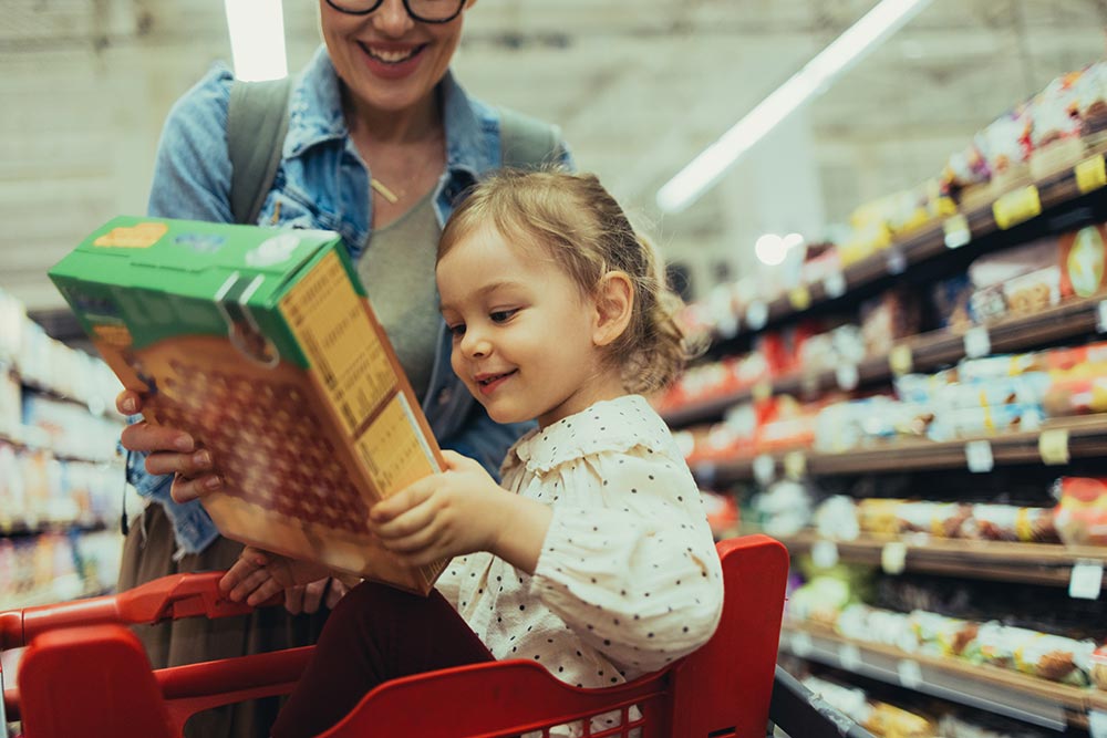 A mother hands a child in her trolley a box of cereal.