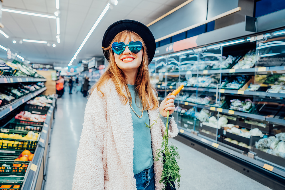 A woman stands in the middle of a supermarket aisle, looking at camera and smiling. She is holding a carrot. 