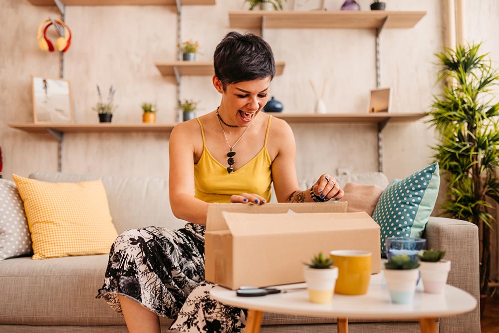 A woman sits on her couch excitedly opening a package that has just been delivered. 