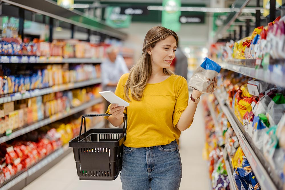 A lady walks down a supermarket aisle, reading the label of a packet she is holding.