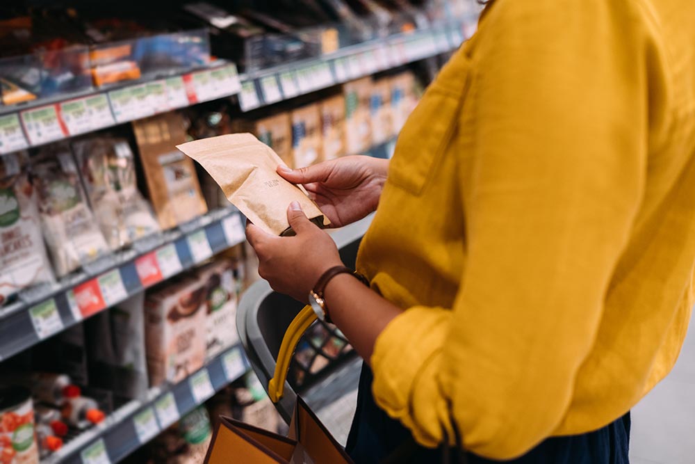 A person holds an unknown product in the baking section of the supermarket. Cake mixes and packets of various flours can be seen in the background.