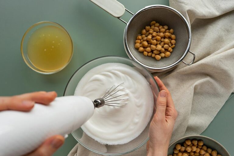 Overhead view of hands using an electric beater to whip up aquafaba. A bowl of extra aquafaba sits to the side, as does a bowl of drained chickpeas.