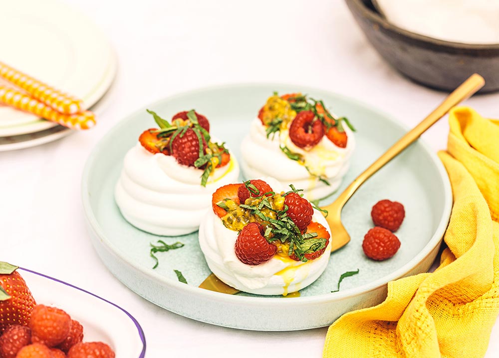 Three mini pavlova cups decorated with passionfruit and raspberries sit in a mint green bowl, with a gold cake server underneath one.