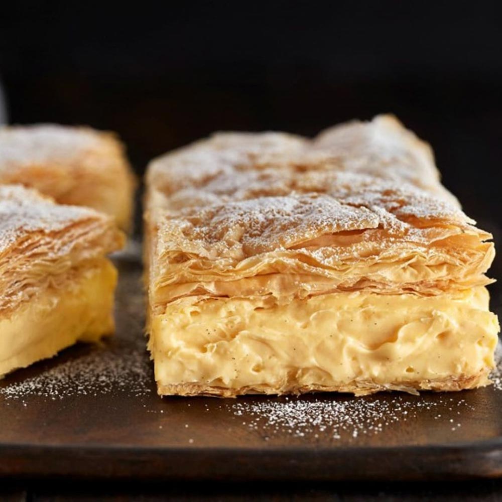 A close-up of vanilla slice squares made with a beautifully flakey pastry. They are topped with powdered sugar. 