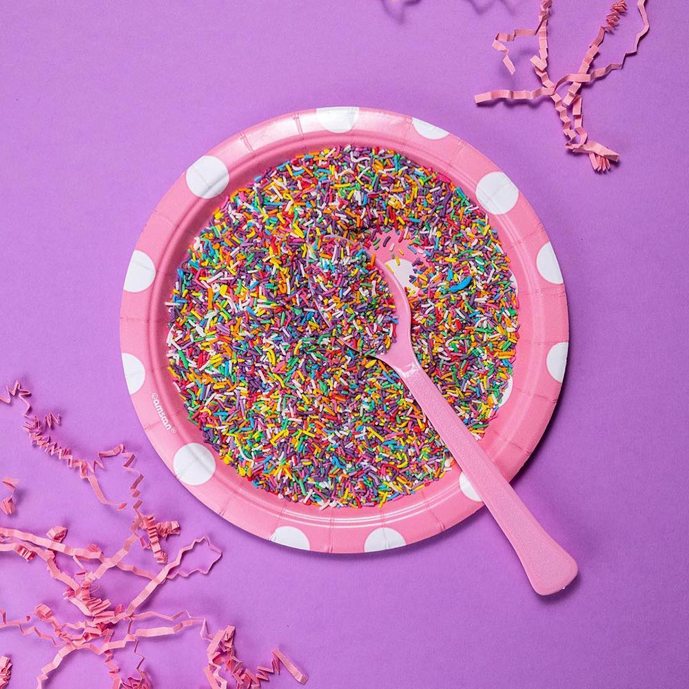 A pink polka-dot plate sits on a purple background. There is a pink spoon on the plate which is covered with sugar sprinkles, creating an explosion of colour.