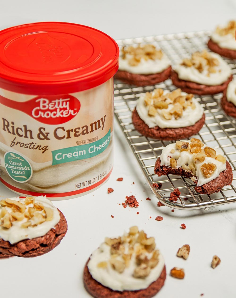 A tub of Betty Corcker ready-made cream cheese frosting sits alongside a tray of cookies that have been topped with the frosting and decorated with walnuts.