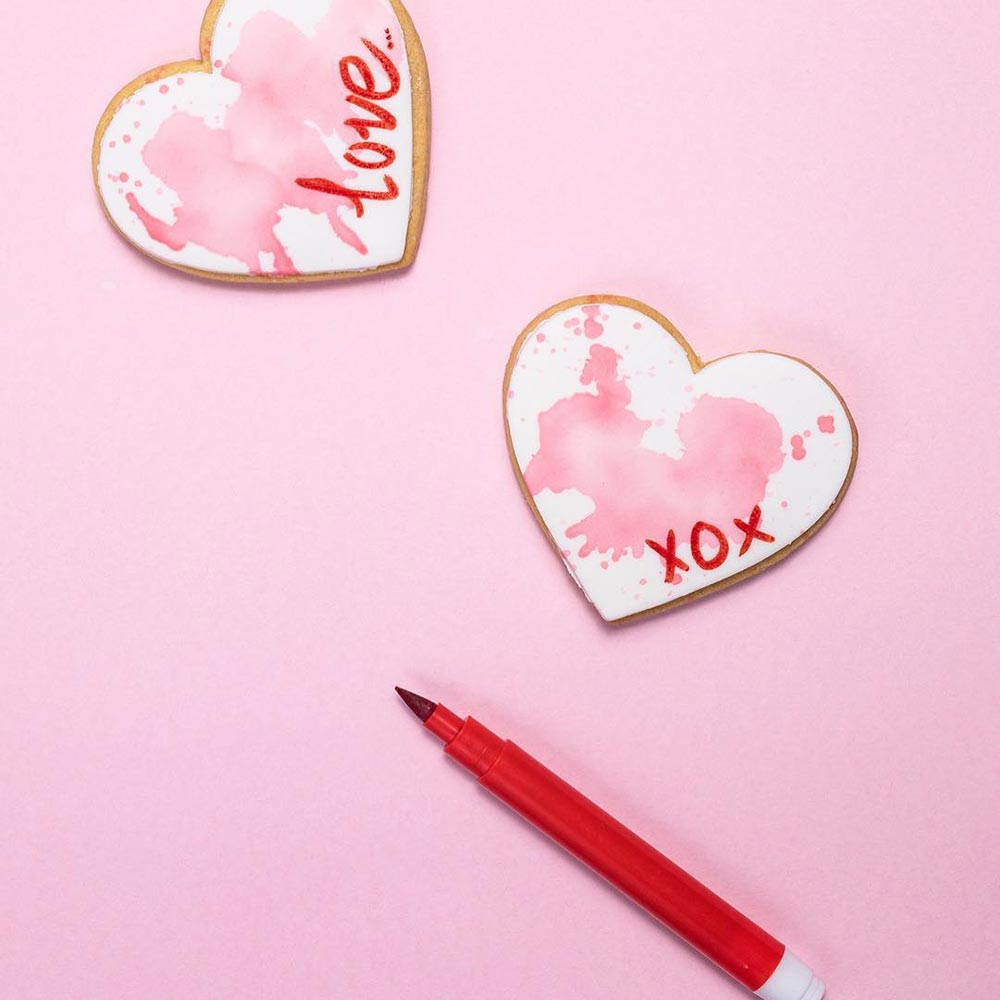 Two heart-shaped cookies are on a pink background. They are iced with white fondant and have 'love' and 'xox' written on them with the edible red marker that sits beside them. A wash has been used to give a watercolour-stained effect.