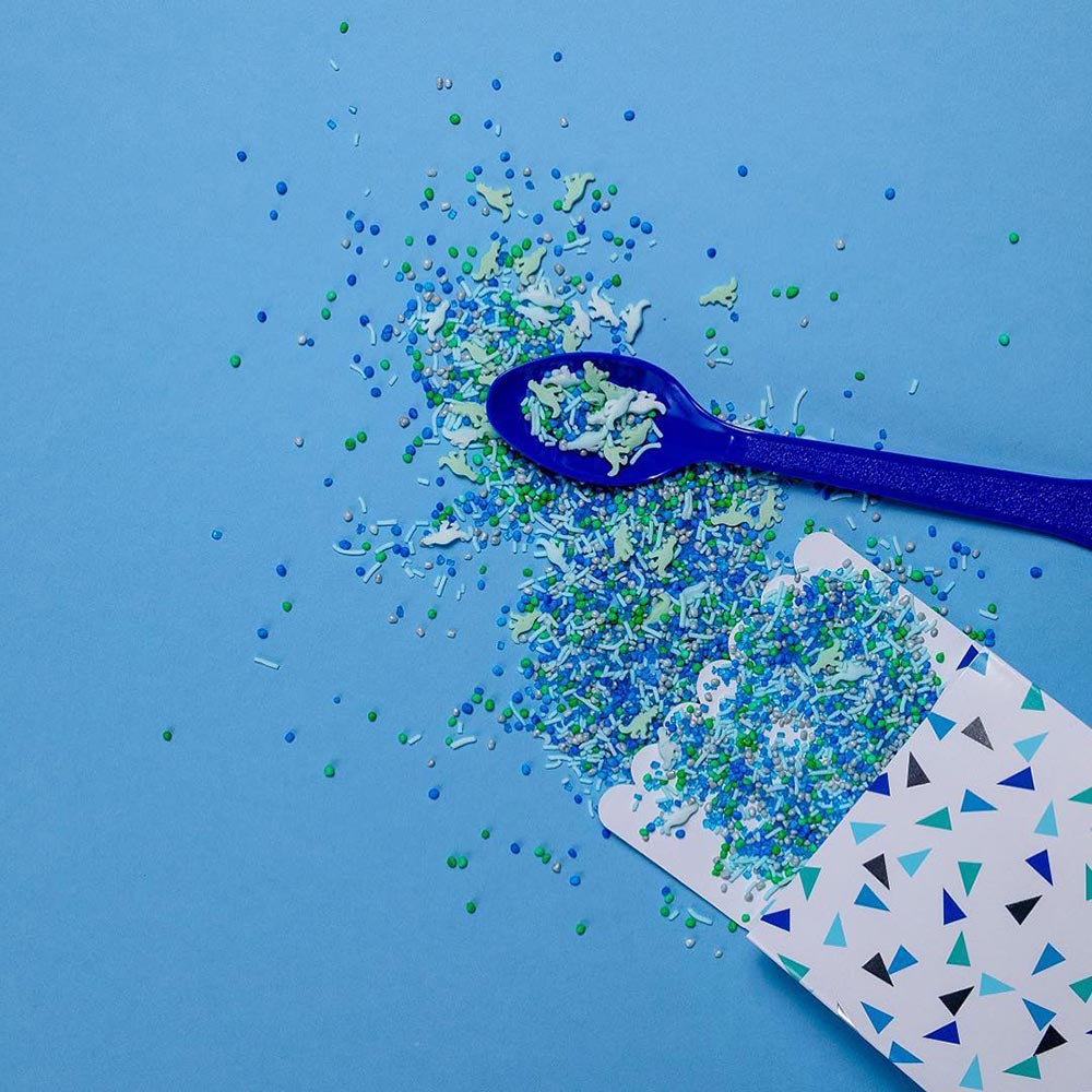 A paper party bag is laid open on a blue background. Out of it, blue and green sprinkles spill out with some in the shape of dinosaurs. A spoon holding them sits nearby.