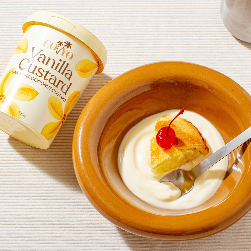 A tub of COYO vanilla custard is laid down on a bench next to a bowl of a sweet cherry topped pie, floating in the creamy custard.