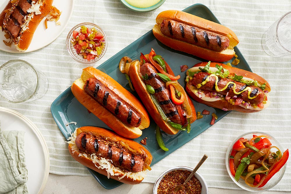 Five hot dogs are on a blue chopping board decorated with different sauces and fillings. There is a range of dishes of other ingredients around to add.