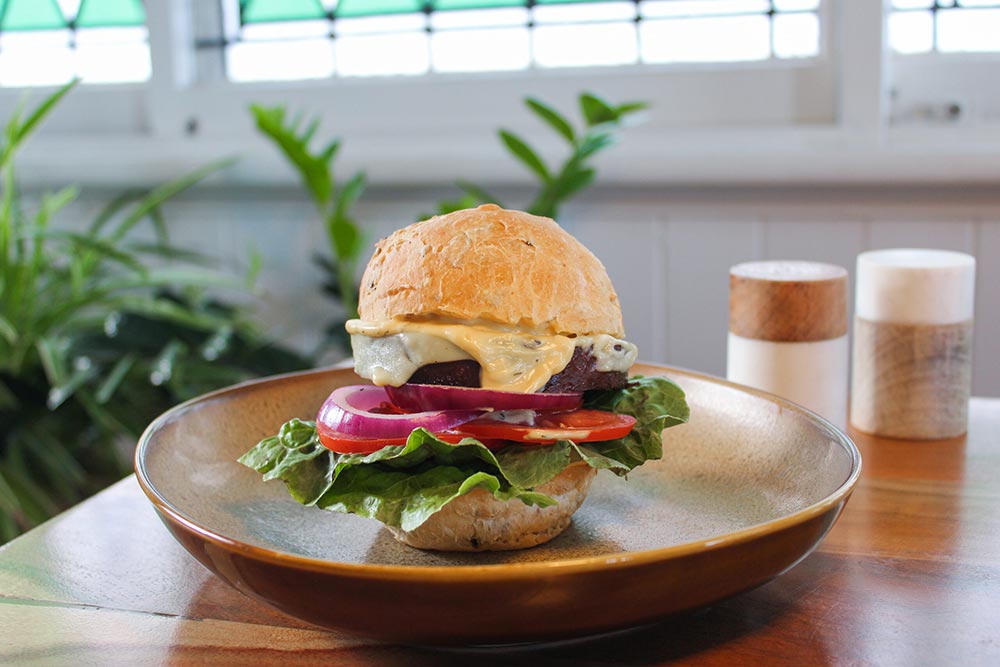 A plant-based cheeseburger with all the trimmings sits on a cafe table. A slice of cheese in the burger is melted, dripping down the side. 