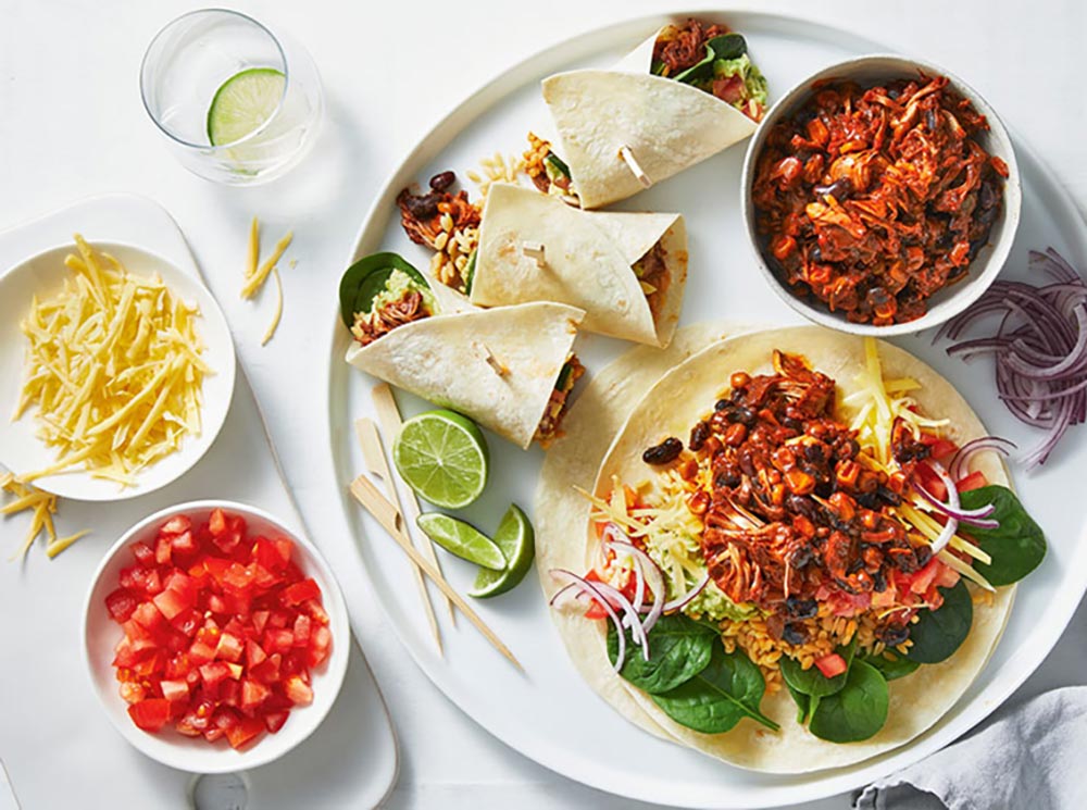 Overhead view of a plate of jackfruit burritos with condiments dishes to the side, filled with jackfruit, plant-based cheese, and diced tomatoes.