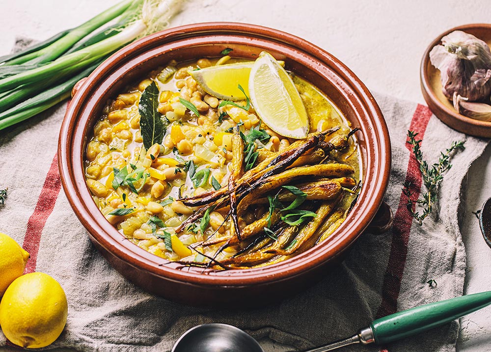 A bowl of stew packed with white beans and topped with lemon wedges, crispy carrots and garnished with spring onion. 