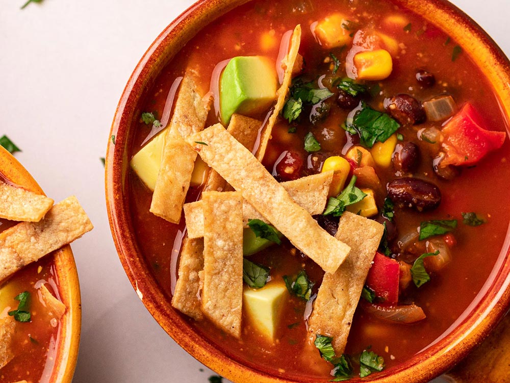 A bowl of tomato-based soup full of kidney beans, avocado, and corn with crispy tortilla strips on top.