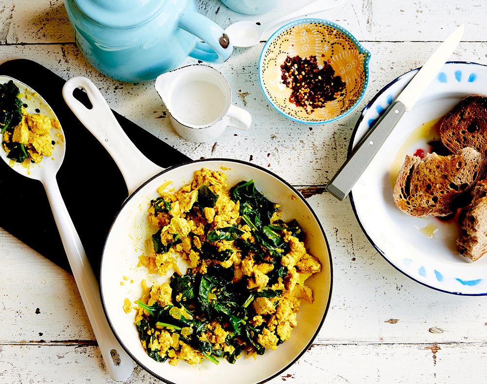 A spinach and tofu scramble is in a white frypan on a table. A plate of toast is nearby, ready to self-serve.