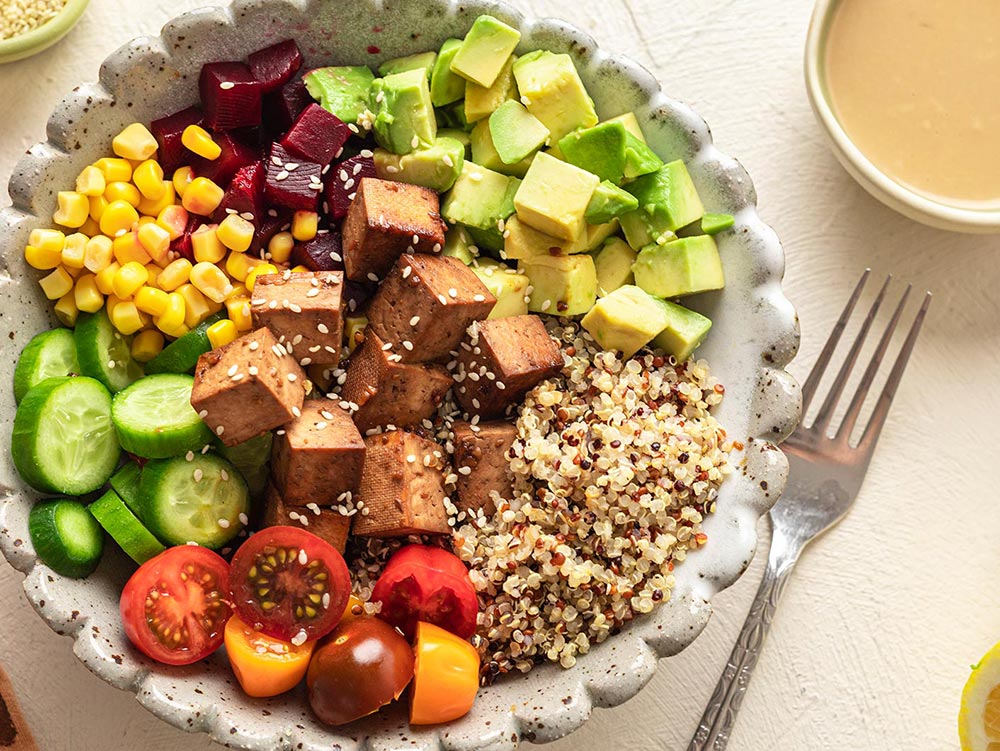 A variety-packed poke bowl with quinoa, cubed avocado, cherry tomatoes, corn, diced beetroot, sliced cucumber, and crispy marinated tofu cubes,