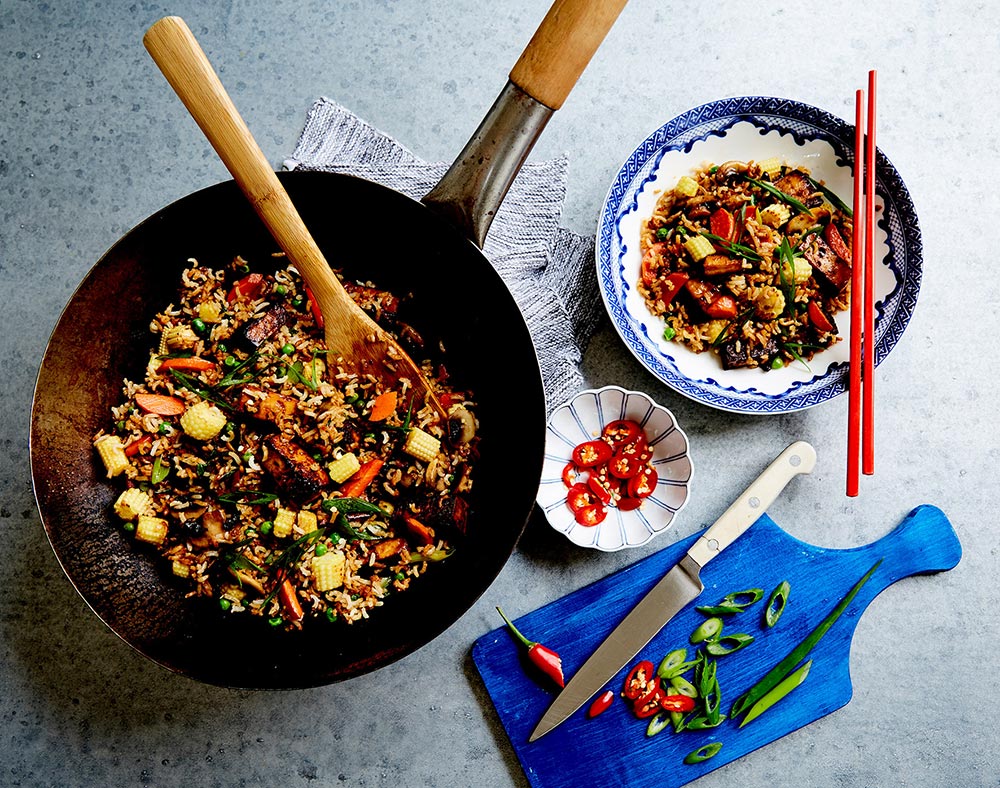 A small wok is filled with colourful fried rice. A bowl of the rice is ready to serve next to it, while a chopping board with fresh chilli and spring onion sits nearby. 