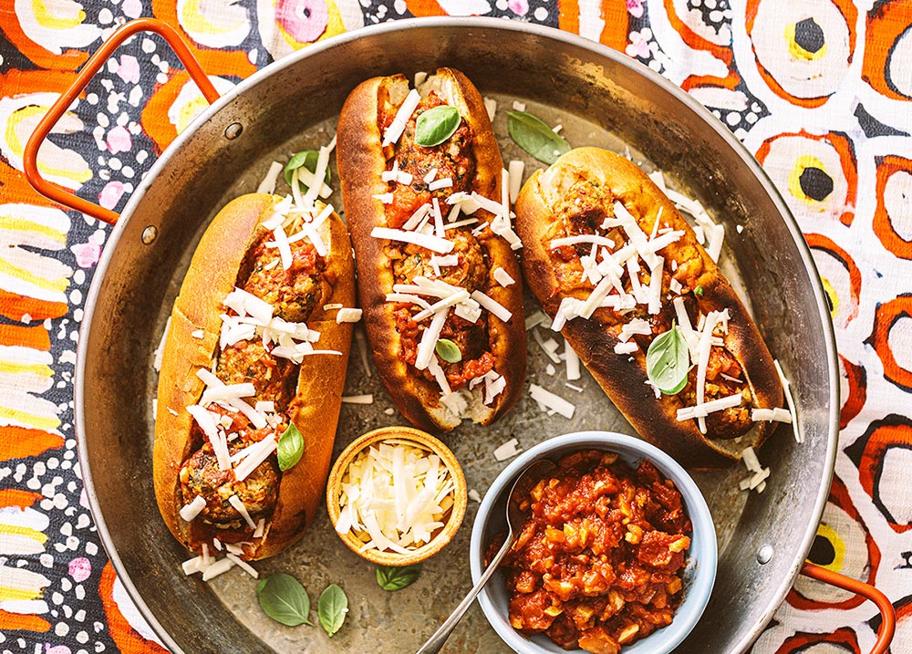 An overhead view of a pan with three toasted plant-based meatball subs. They are topped with sauce and cheese, with more of both in little bowls to the side.