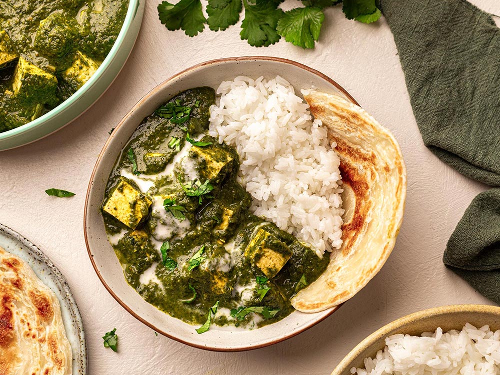 A bowl of palak 'paneer', the paneer made from tofu. The curry is in a rich green spinach gravy with a aside of rice and roti. 