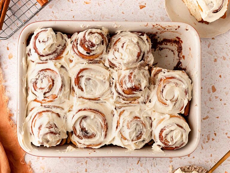 An overhead view of a tray of freshly iced cinnamon scrolls. One has been taken from the tray and is sitting on a plate just in shot.