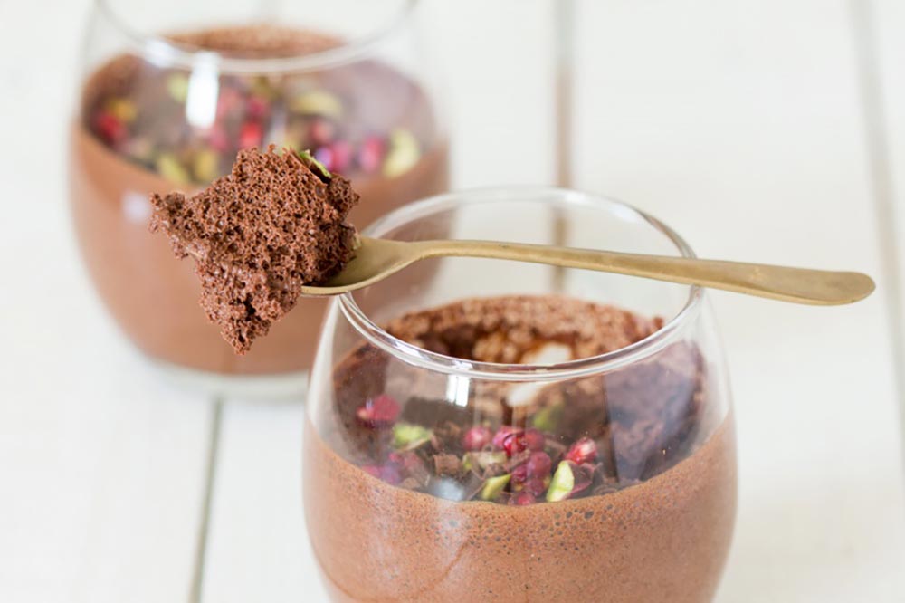 Two dessert tumblers of chocolate mousse sit on a wooden tabletop. They're topped with pomegranate seeds and pistachios. A spoon sit on one of the tumblers with a scoop of mousse on it, showing how light and airy it is.