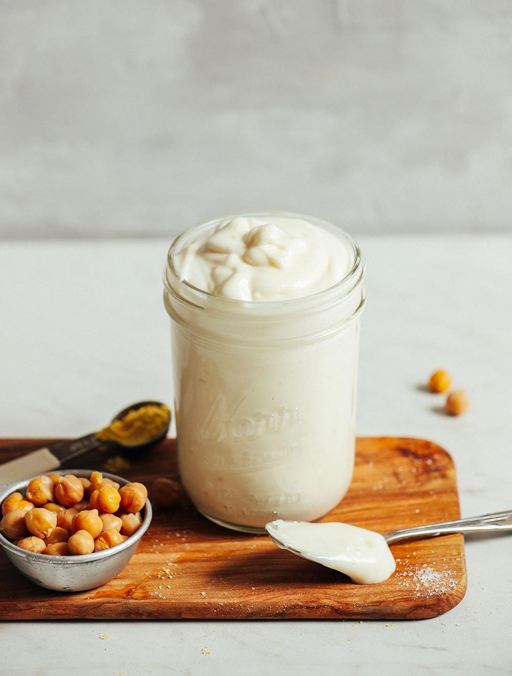 A jar sits on a chopping board, filled to the brim with mayonnaise. Chickpeas sit to the side in a small dish, with a spoonful of mayo nearby.