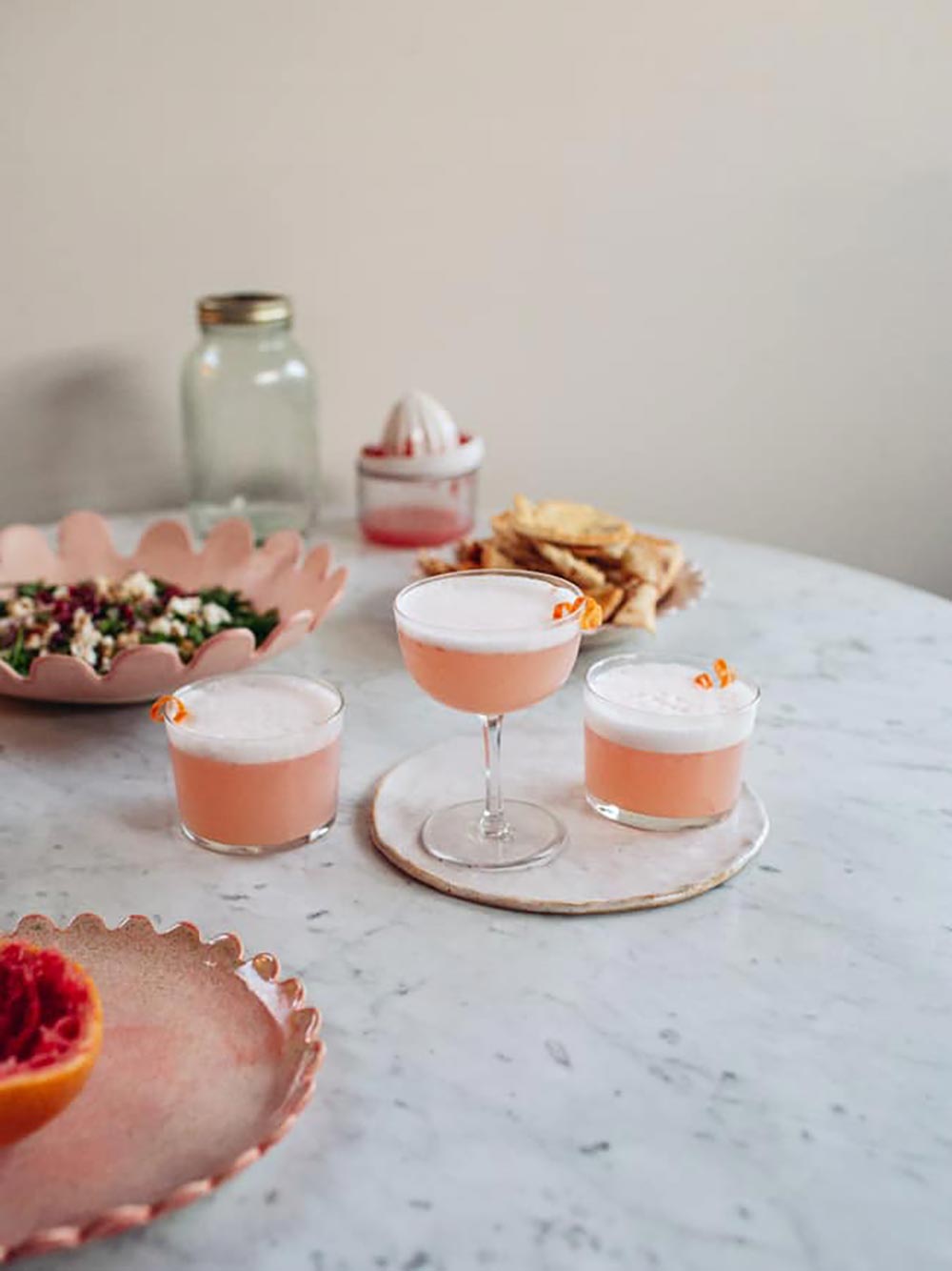 Three grapefruit gin sour cocktails sit on a table surrounded by snacks. They have a creamy head on them, thanks to the use of aquafaba. 