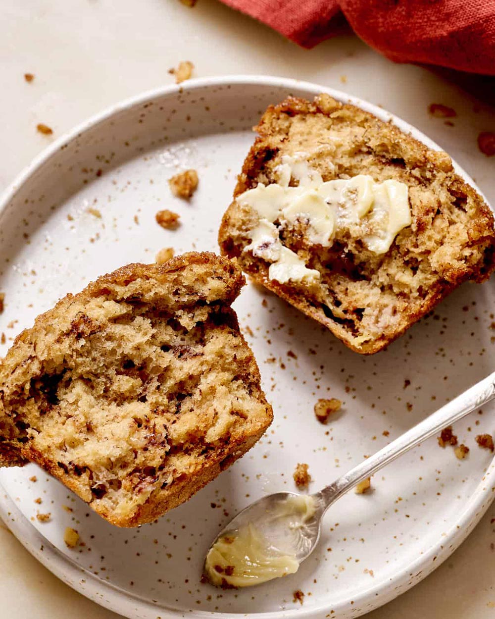 A banana muffin cut in half is on a plate. One half is slathered with butter, the other facing the camera to show the moist cakey texture.