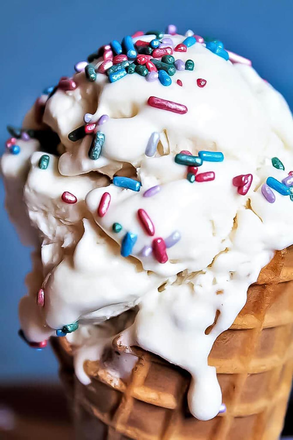 A close-up of an ice cream cone with vanilla ice cream dribbling down the side. It's decorated with colourful metallic pink and blue sprinkles.