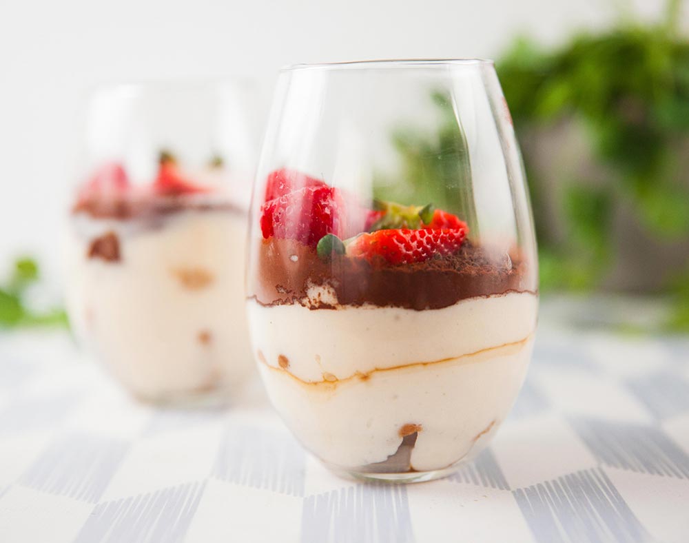 Two glass dessert tumblers are filled with tiramisu, showing the layers of cream, cocoa, and cake. They're topped with strawberries. 