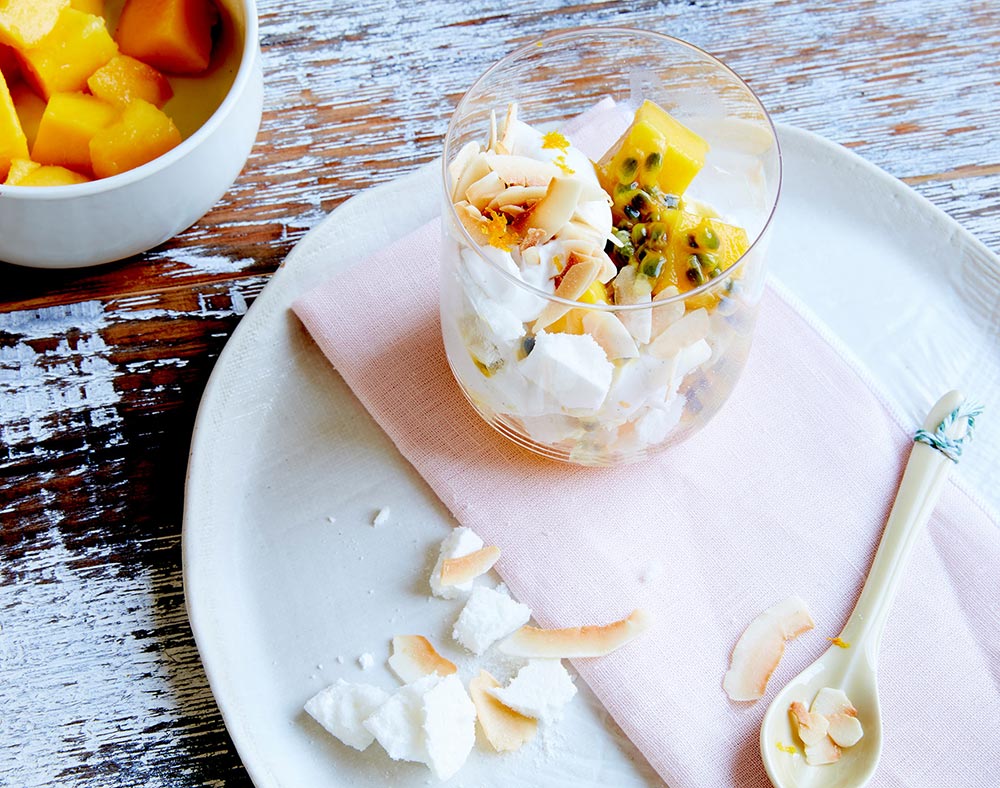 Overhead view of a glass dessert tumbler filled with Eton Mess - crushed meringue, yoghurt, mango cubes, passionfruit and toasted coconut.