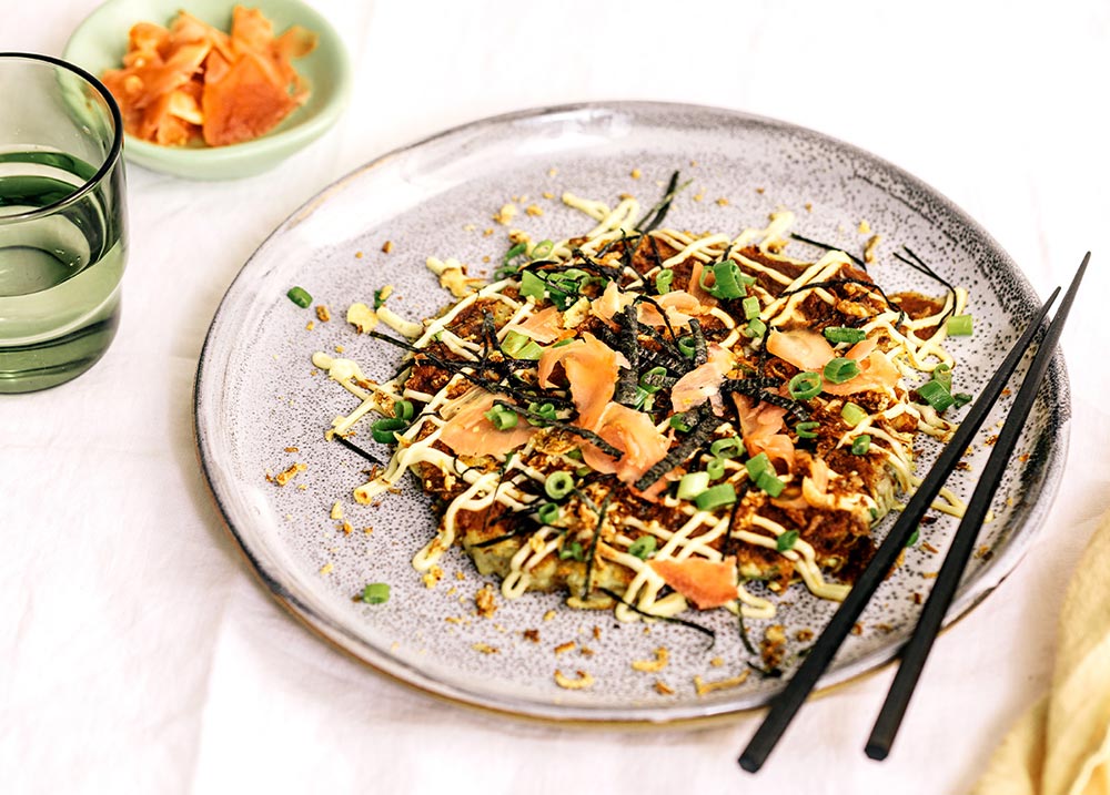 A Japanese pancake is served on a plate at a table setting. It's topped with sauces, pickled ginger, spring onions and herbs.