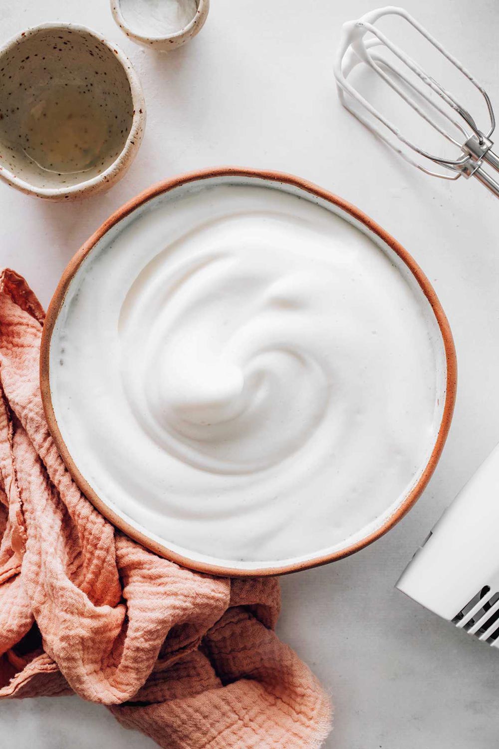 An overhead view of a bowl full of whipped aquafaba cream. A hand mixer is on the bench to one side with a tea towel. The cream has formed stiff peaks.