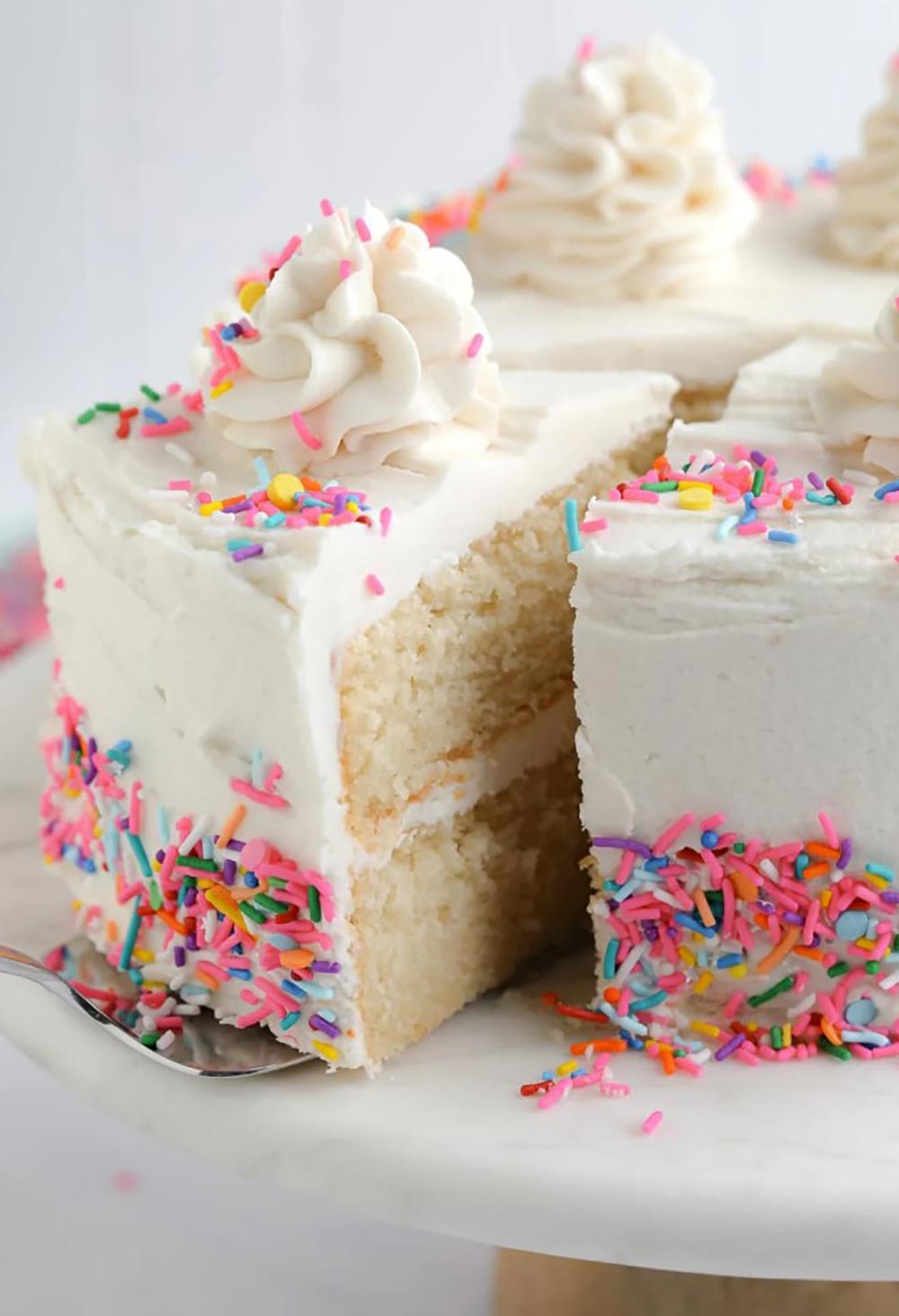 A slice of vanilla cake is being removed on a cake server. The texture is very moist, light, and fluffy. It's decorated beautifully with a white buttercream layer and frosting, decorated in brightly coloured sprinkles. 