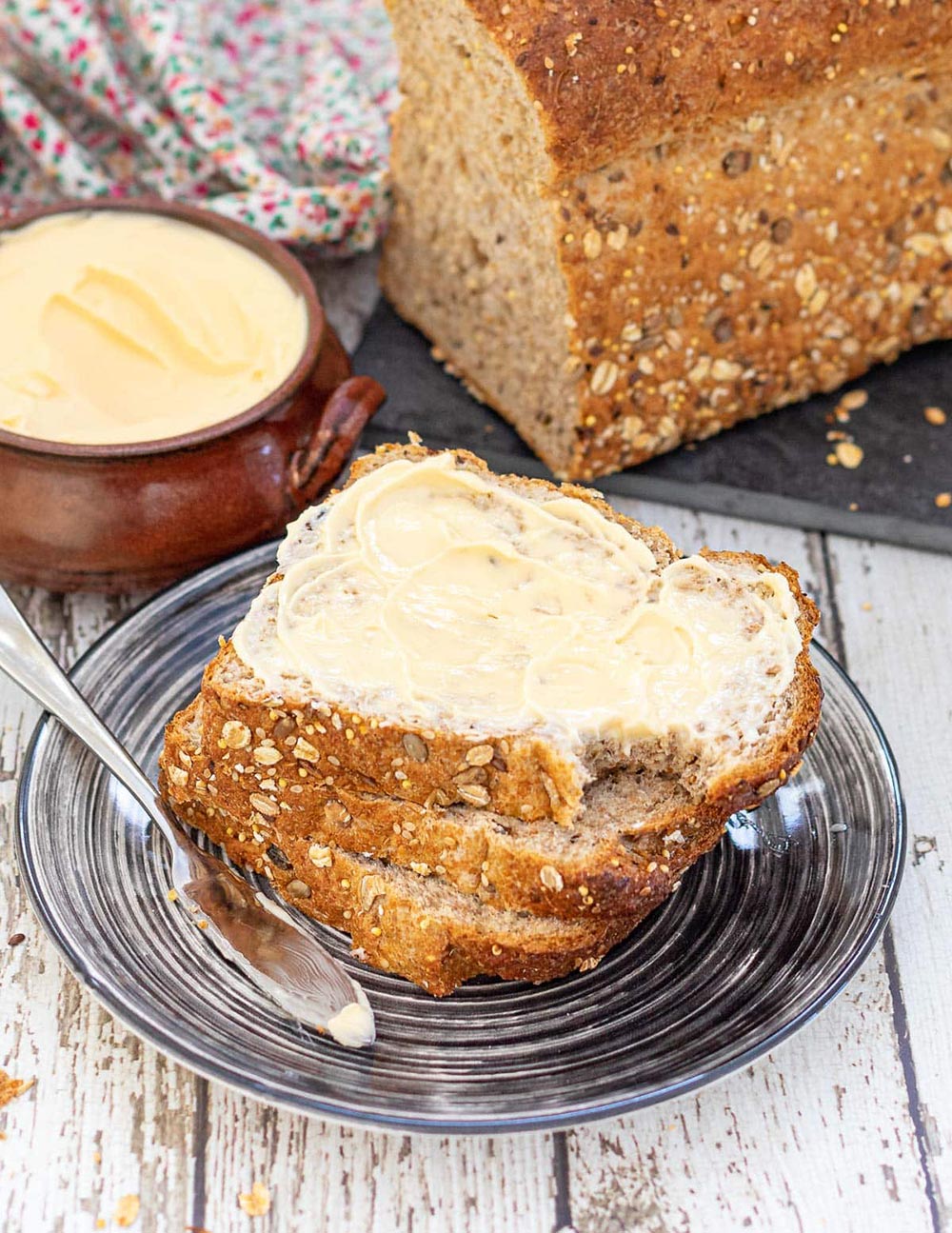 A plate is piled high with sliced bread, the top slice slathered with the homemade vegan butter that sits in a dish behind it. 