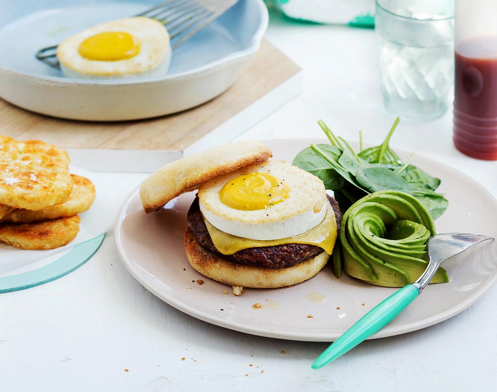 An English muffin with a vegan egg, cheese, and a plant-based beef patty sits on a plate, with a side of spinach and avocado. Hash brown are in a pile on a plate to the side. 