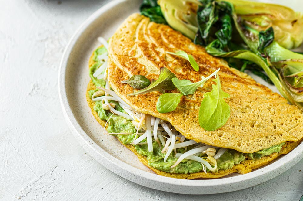 An omelette sits on a plate folded over. It's filled with avocado and bean shoots, topped with fresh greens. Asian greens are served on the side. 