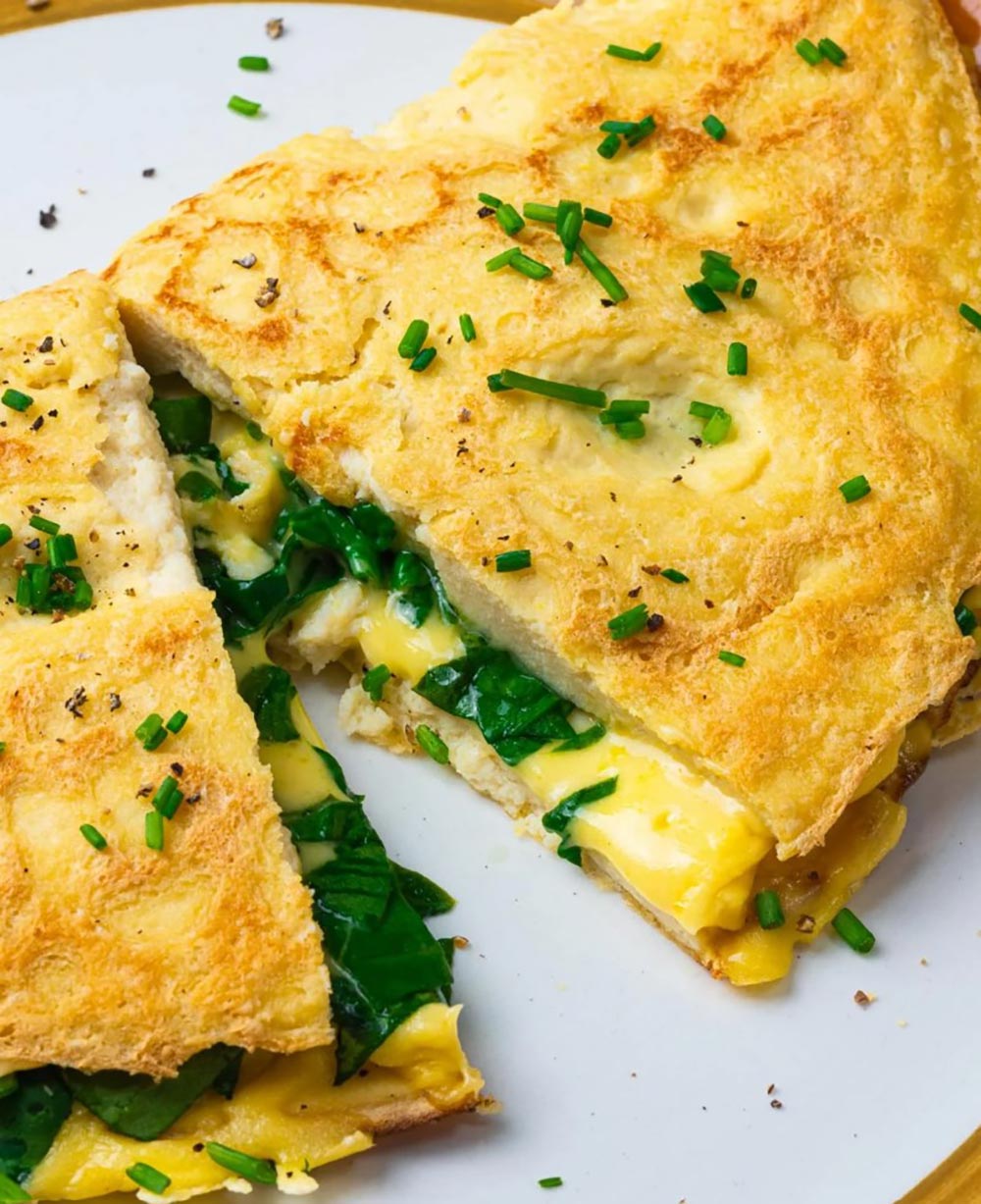 A close-up view of an egg-free omelette garnished with fresh greens. It's been cut in half to show it's full of melty vegan cheese and spinach. 