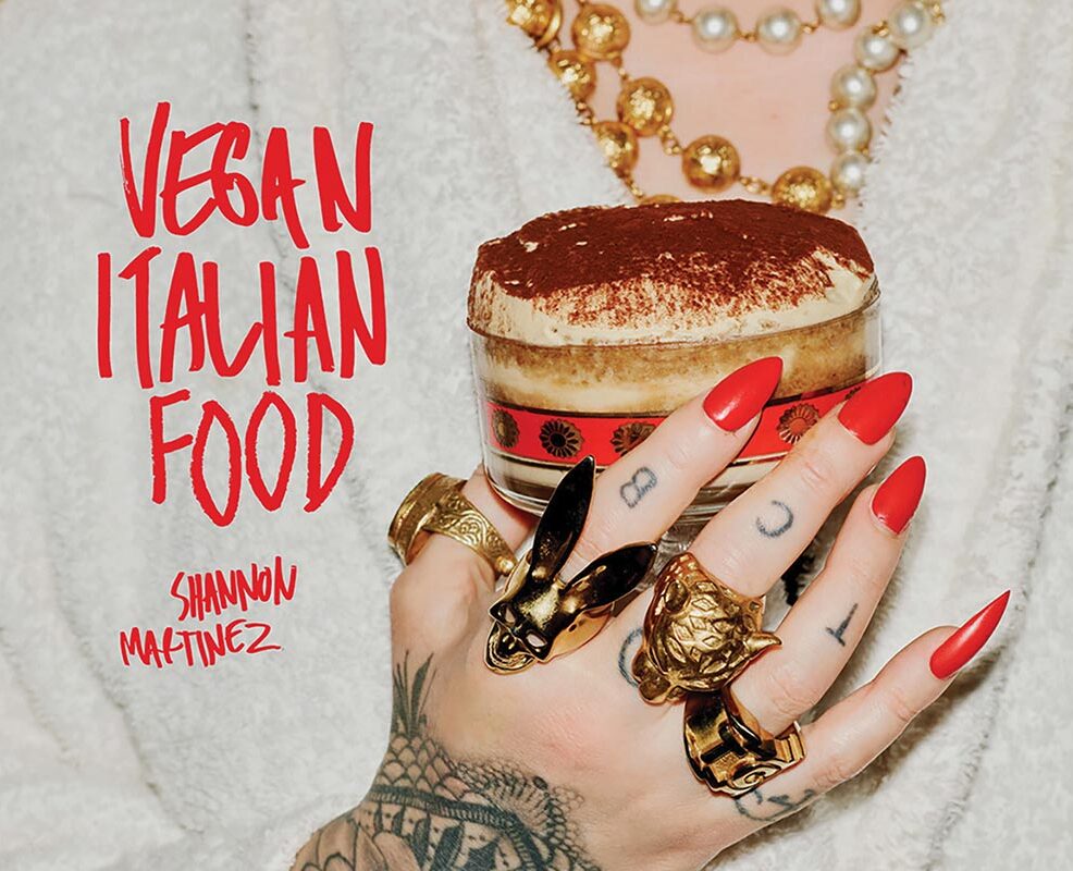 The cover of Vegan Italian Food by Shannon Martinez. Shannon is clutching an Italian dessert to her chest. Her tattooed hand is adorned with numerous gold rings and freshly painted red nails.