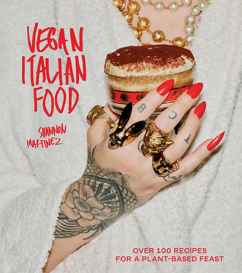 The cover of Vegan Italian Food by Shannon Martinez. Shannon is clutching an Italian dessert to her chest. Her tattooed hand is adorned with numerous gold rings and freshly painted red nails.